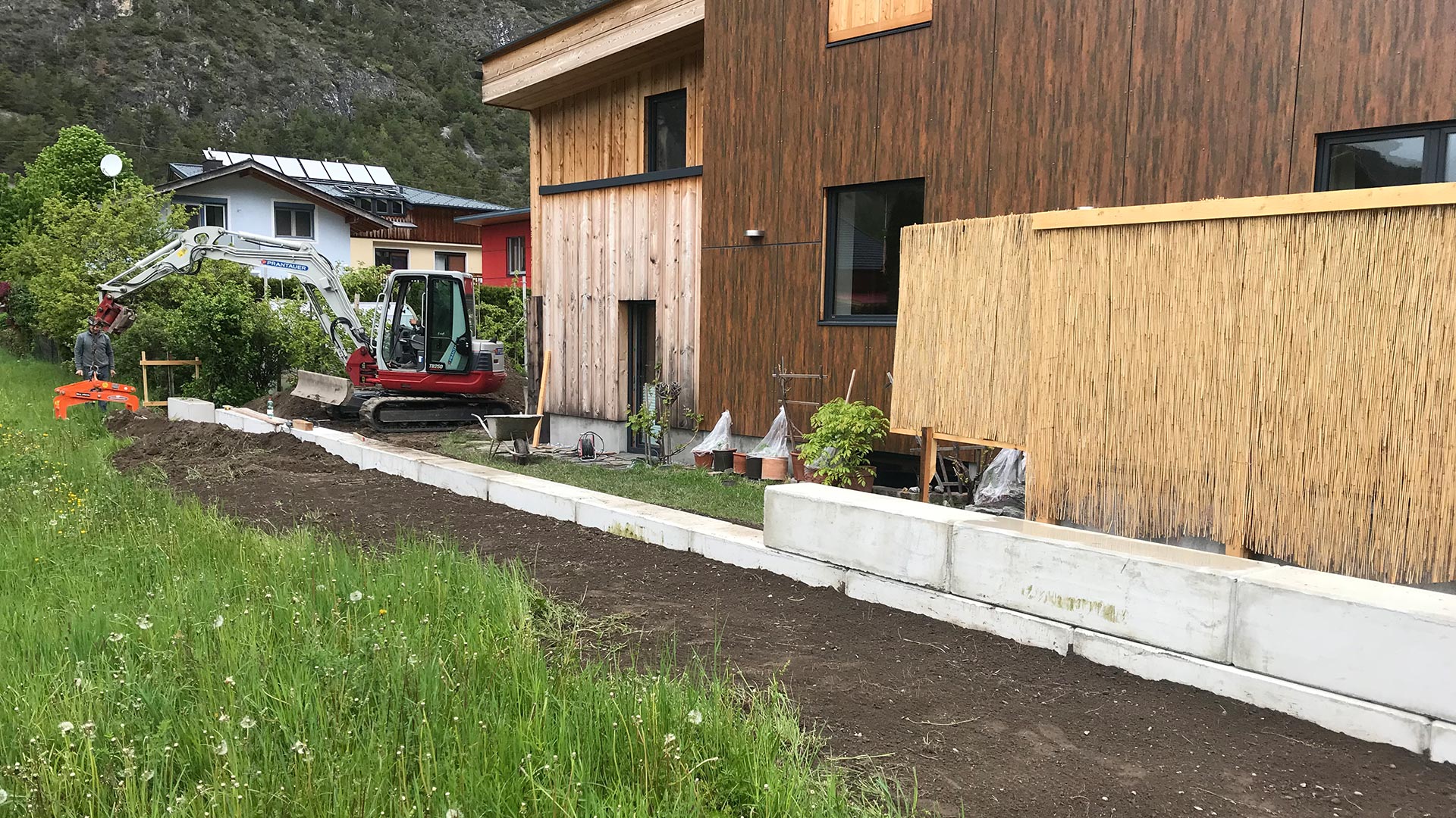 Mauer aus Betonfertigteilen mit Bagger und Einfamilienhaus